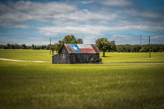 Old Rustic Barn With Texas Flag Painted on Roof With Old Wagon Nearby In Green Field in Rural Town in Texas