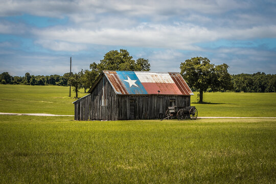 Old Rustic Barn With Texas Flag Painted on Roof With Old Wagon Nearby In Green Field in Rural Town in Texas