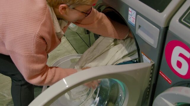 Woman in pink sweater places laundry basket near washing machine, loading white clothes into the drum while standing in a self-service laundromat with modern appliances
