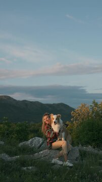 A girl in a red shirt is stroking and hugging a border collie, sitting on a rock at the top of a mountain. Sunset on a journey and friendship with animals