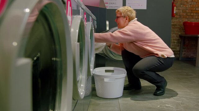 Senior woman in pink sweater loading laundry into washing machine at self-service laundromat, with white basket and modern appliances in view