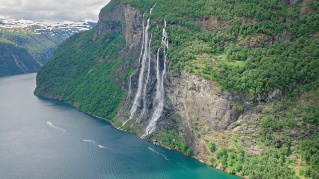 Drone flies closer to the Seven Sisters waterfall cascading down a vertical rock face. Iconic Norwegian nature unfolds above the calm waters of Geirangerfjord.
