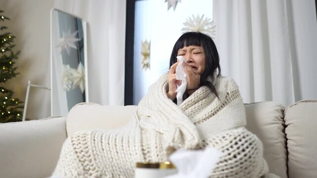 sick young Asian woman covered with blanket suffers from cold and runny nose at home, the unhappy girl wipes her nose with tissues and shows flu symptoms