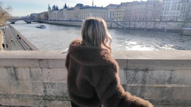 A woman in a fur coat is seen waving on a bridge over the Seine River in Paris France She is taking candid selfies for social media on a sunny day with a cityscape background The shot is handheld.
