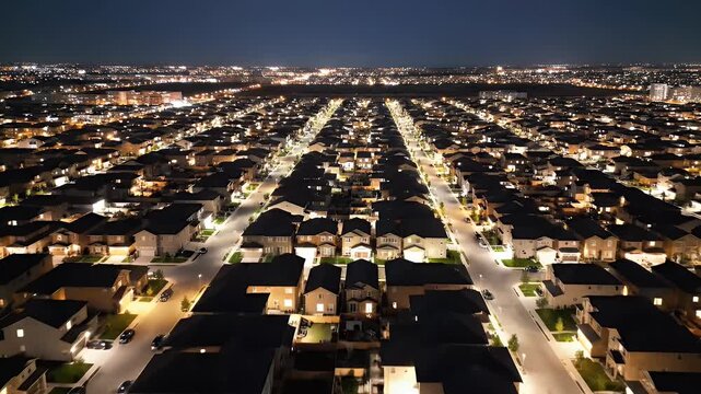 Aerial night view of suburban homes, lit by streetlights, with city lights in the distance