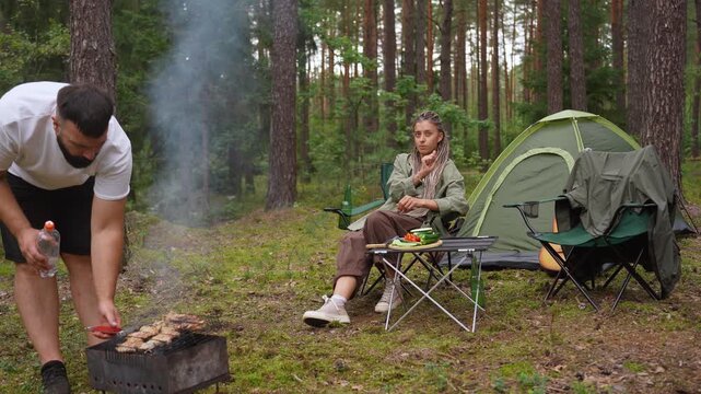 Young man grilling meat on a barbecue while his girlfriend sits nearby at a campsite with a tent. The couple is enjoying a peaceful picnic and outdoor recreation in a beautiful pine forest