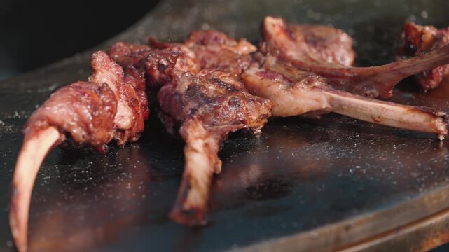 A close-up, selective focus shot of juicy lamb chops cooking on a hot barbecue griddle. The meat is browned and sizzling, with visible bone. Warm lighting enhances the appetizing, savory mood