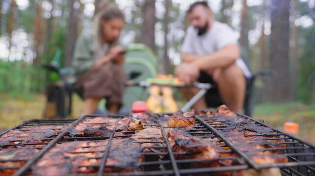 Delicious pork meat grilling on a barbecue rack over a campfire during a picnic in the woods, with a blurry couple of friends relaxing in the background and smoke rising from the hot charcoal