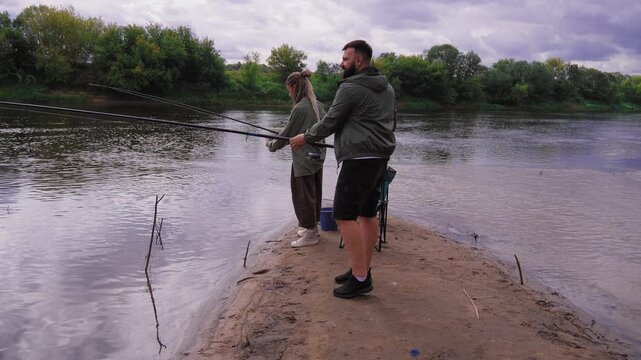 Millennial man and woman enjoying a relaxing day of fishing together, standing on a sandy riverbank with their rods and reel, sharing a peaceful outdoor hobby during the weekend