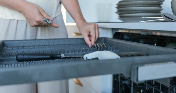 Woman placing cutlery into dishwasher upper rack in modern kitchen