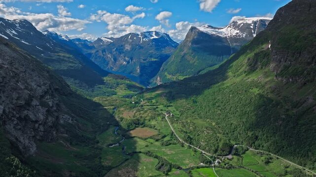 Drone shot gliding deeper into a lush green valley, with farms and narrow roads scattered across the valley floor. Snow-dusted peaks rise in the distance under clear summer skies.