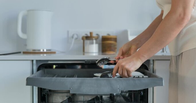 Woman placing cutlery into dishwasher upper rack in modern kitchen