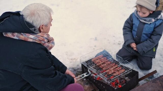 Gray haired grandfather and child by grill roasting sausages on charcoal brazier, bundled in winter coats and scarves on snowy ground. Hands warmed over glowing embers, smoke. Family outdoor barbecue.
