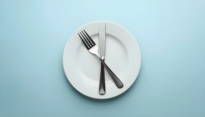 A top down view of a white plate with a fork and knife on a light blue colored surface in a studio
