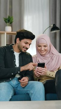 Attractive bearded 30-aged arabian man in casual clothes sitting with his lovely cheerful girlfriend in headscarf on soft couch in contemporary living-room and use smartphone to revision joint photos
