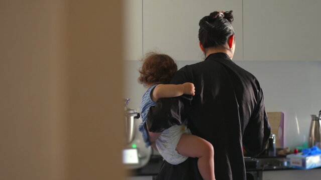 Woman holding toddler in modern kitchen, standing near appliances with back turned, caring for child while engaged in household task