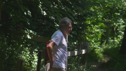 Older man breathing deeply while resting by wooden fence along forest path, pausing after summer hike and leaning on railing surrounded by trees and dense greenery