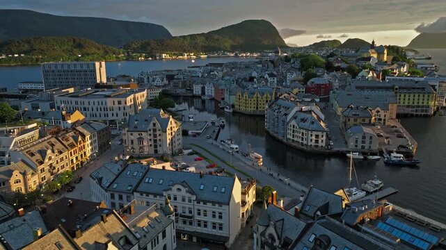 erial drone view of Alesund, Norway over the inner harbor during golden hour sunset.The norwegian scene shows canals, curved waterfront buildings, bridges, docked boats, and dense city blocks.