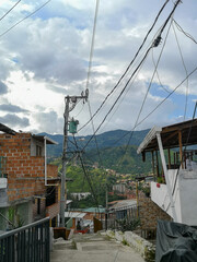 A steep residential sidewalk in the mountain suburbs of Medell&iacute;n with overhead power lines. Brick houses and white walls overlook green valleys and distant hills beneath a partly cloudy sky.