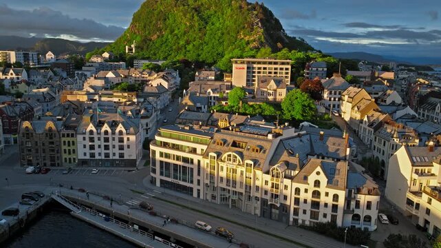 Aerial drone view of Alesund, Norway with a steep green hill rising behind the city at golden hour sunset.The Norwegian scene shows clustered buildings, harbours, water, and quay lines.