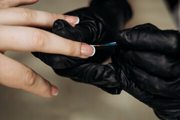 A technician wearing black gloves carefully applies a white French manicure to a nail with a thin brush. Close-up, professional manicure.