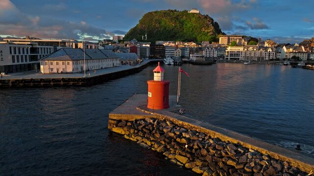 Aerial drone view of Alesund, Norway featuring a red lighthouse, rocky pier, and surrounding water during golden hour sunset. From above, navigation structures and shoreline define the Norwegian port.