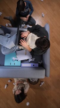 Vertical video father juggling remote work on laptop while two kids create chaos with crumpled paper in a cluttered living room, stressed and distracted