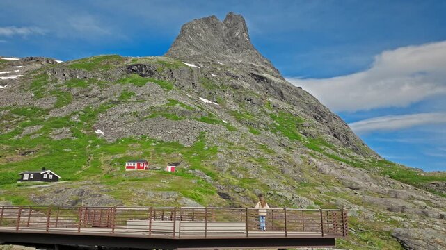 A tourist stands on the Trollstigen viewing platform, facing a steep rocky slope with green patches and scattered cabins. Along the mountainside, rugged rock formations and open sky dominate the lands