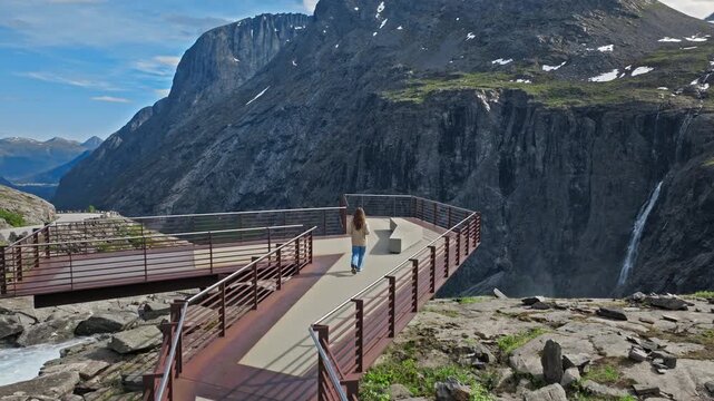 A tourist moves along the curved Trollstigen viewing deck overlooking a dramatic cliff drop. Beyond Trollstigen, dark rock walls and deep valleys stretch into the distance.
