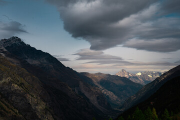 Fototapeta premium Dramatic mountain valley landscape under a moody cloudy sky in the Swiss Alps. Dark mountain slopes framing a distant sunlit peak at dusk. Cinematic alpine scenery near Zermatt, Switzerland.