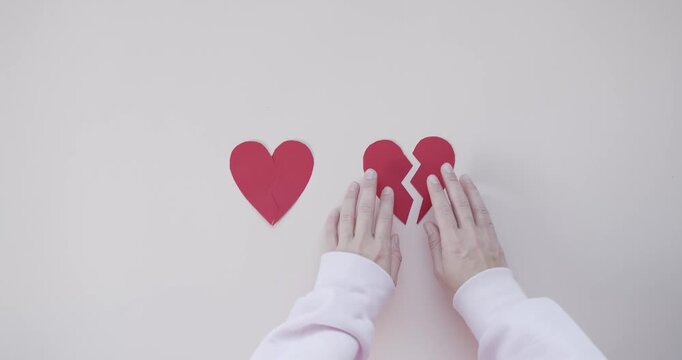 Hands Touching Two Broken Red Paper Hearts on White Surface