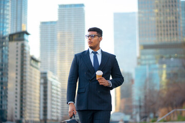 Fototapeta premium A black man in a suit stands in an urban area holding a coffee cup. He looks around as tall buildings surround him. It is a sunny day and he seems focused on his day ahead.
