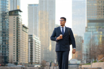 Fototapeta premium A black man in a suit walks along a city street holding a coffee cup. Skyscrapers surround him as he moves forward. The scene shows a busy urban environment in daylight.