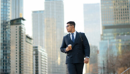 A businessman walks along a city street holding a cup of coffee. Tall buildings surround him as he...