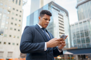 Fototapeta premium A black man in a suit stands outside tall buildings in a city. He is focused on his smartphone and seems to be engaged in an important task. The environment is urban and busy.