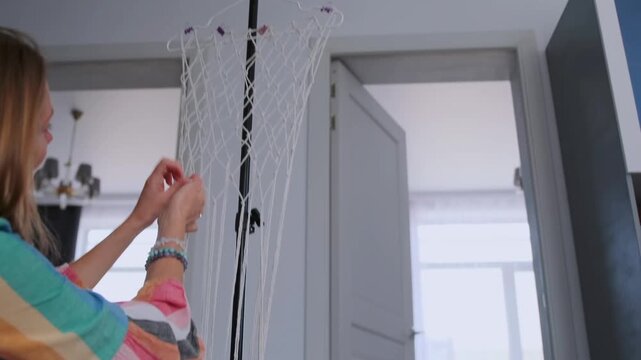 Young woman in colorful dress weaving macrame on the floor -  smiling and inspired in bright living room. Handmade craft and cozy lifestyle.