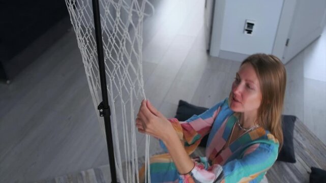 Young woman in bright dress weaving macrame on the floor of a light living room -  smiling and chatting with her friend. Cozy lifestyle and handmade craft. Top view.