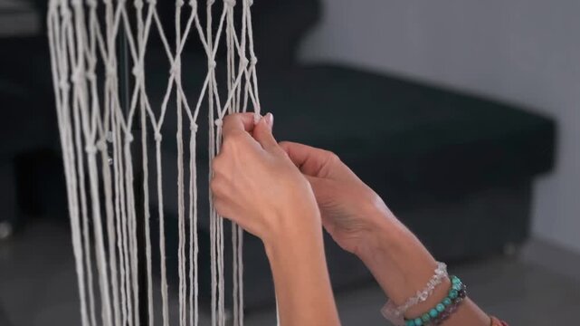 Close up of woman hands with bracelets tying knots in macrame threads. Handmade process -  boho craft and creative home hobby.