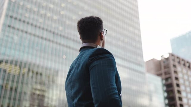 A man stands outside in a suit with headphones around his neck. He looks up at the tall buildings in an urban setting. The sky is bright, and he appears to be deep in thought.