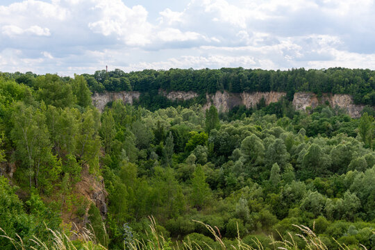 Summer landscape of an abandoned limestone quarry near gora kraka mountain in Luban, featuring steep rocky walls, dense deciduous forest, and a wide green valley under a partly cloudy sky.