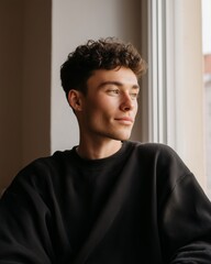 Fototapeta premium Young man with curly hair wearing a black sweatshirt sitting by a window, gazing thoughtfully outside, natural light illuminating his face and the interior space