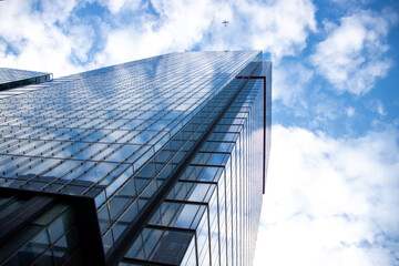 Obraz premium Glass skyscraper reflecting clouds under blue sky with airplane overhead, viewed from a low angle