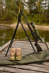 hunting boots on a wooden pier by calm water, with a shotgun on a stand equipped with a scope. outdoor hunting equipment arranged near the lake in a quiet rural nature setting.