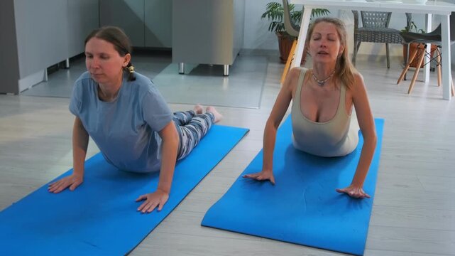 Two women practicing Bhujangasana -  Cobra Pose -  during yoga training at home. Exercise focused on strengthening the back -  improving posture and supporting spine health.