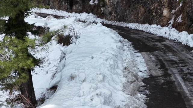 Snow accumulation alongside a winding road with patches of asphalt visible and greenery in the background