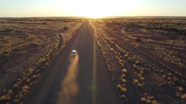 Aerial drone view of a white car driving on a dusty gravel road in Namibia at sunset with beautiful lens flare and golden light