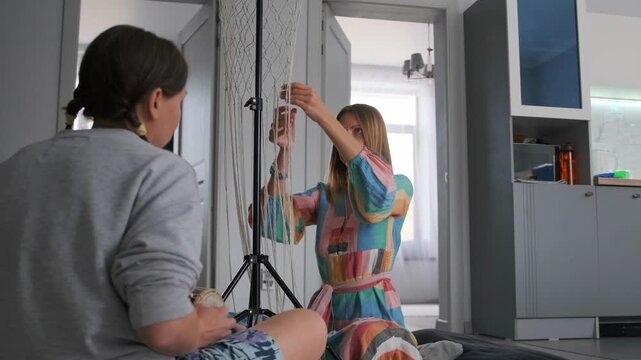Female leisure at home. One woman weaves macrame while her friend plays a drum beside her -  enjoying creative time and friendship in a cozy atmosphere.