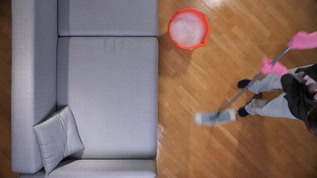 Person mopping parquet floor near modern gray sofa, wearing gloves and using bucket, top down view showing household cleaning routine, tidy living room and hardwood maintenance
