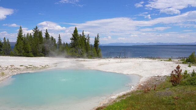 A wide shot of vivid blue geothermal water formed by a geyser with Yellowstone Lake visible in the background at Yellowstone National Park, Wyoming.