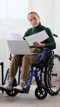A disabled woman in a wheelchair works from home, holding papers and using a laptop. Vertical video.